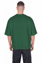 A man viewed from the back wearing an oversized dark green t-shirt and black cargo pants, set against a plain white background.
