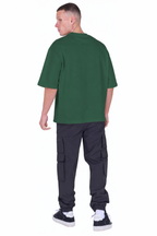 A man shown from the back wearing an oversized dark green t-shirt, dark cargo pants, and black and white sneakers, set against a plain white background.