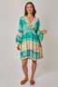 Woman wearing a green and beige patterned dress holding a straw bag on a white background