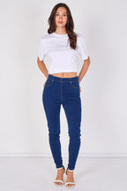 A woman modeling a pair of dark blue skinny jeans with a white cropped t-shirt and white heeled sandals, standing against a plain white studio background.