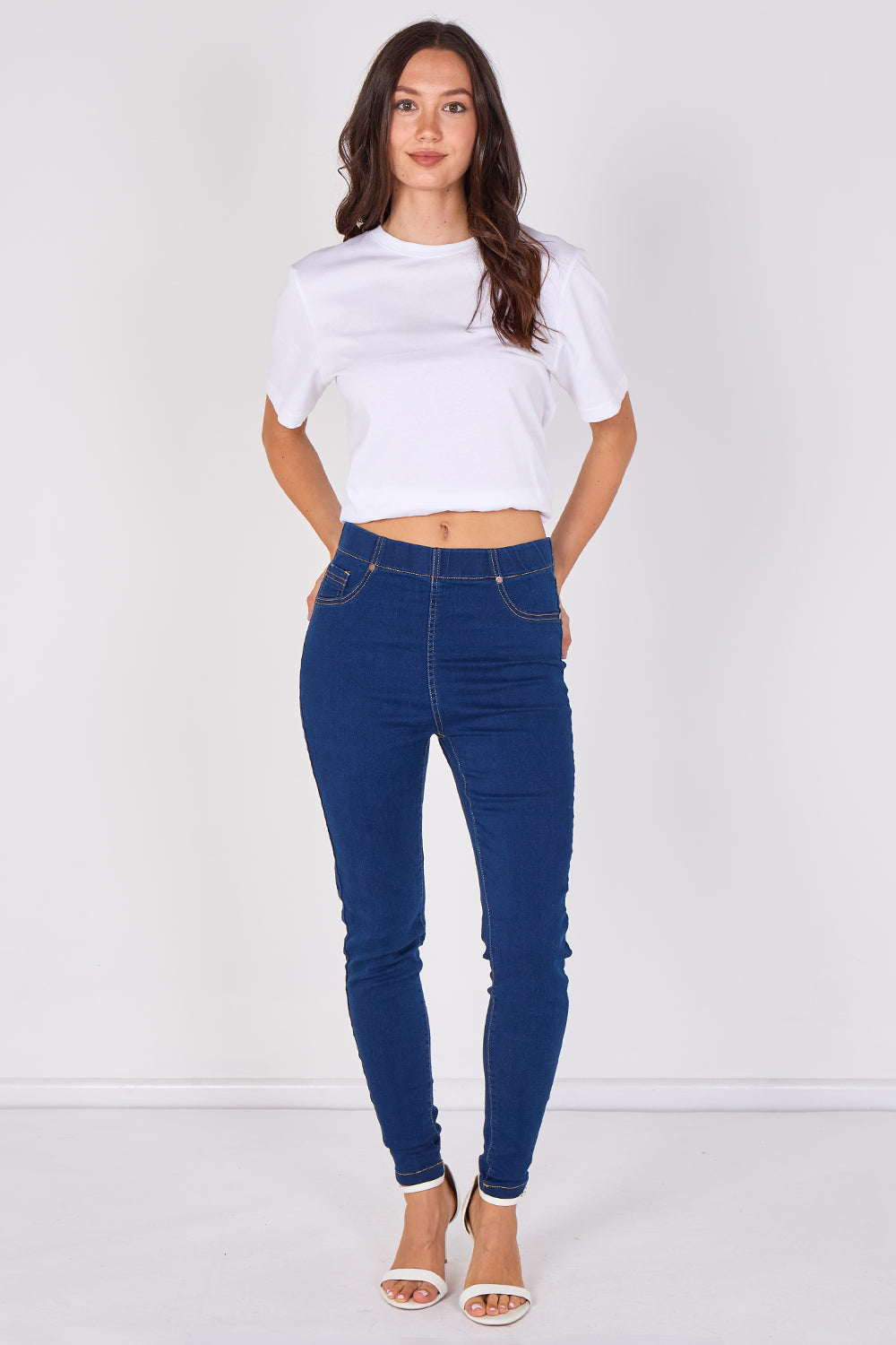A woman modeling a pair of dark blue skinny jeans with a white cropped t-shirt and white heeled sandals, standing against a plain white studio background.