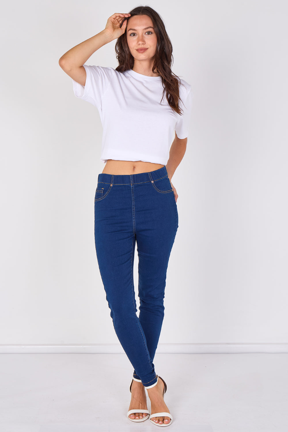 A woman modeling a white cropped short-sleeve t-shirt and dark blue skinny jeans with white heeled sandals, standing against a plain white studio background.