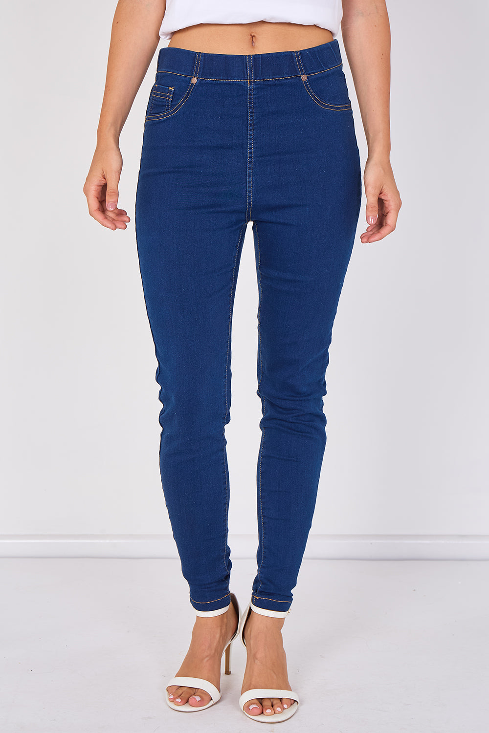 A model wearing dark blue high-waisted skinny jeans with rolled cuffs and white heeled sandals, shown from the waist down against a white studio background.