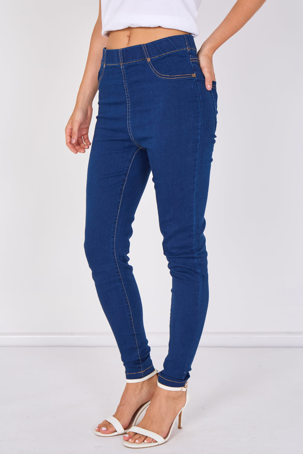A model wearing dark blue high-waisted skinny jeans with white heeled sandals, shown from the waist down against a plain white studio background.