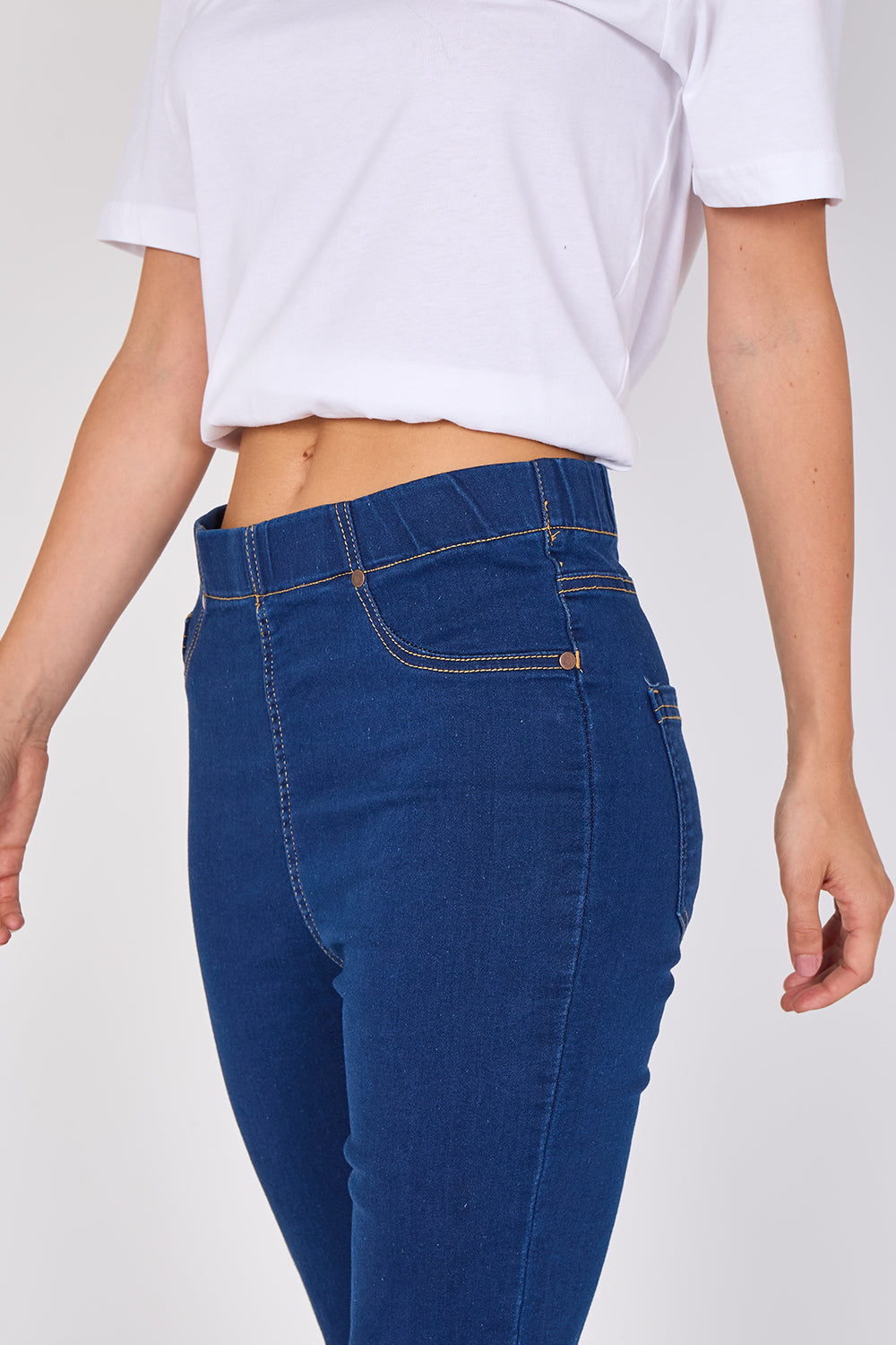 A person wearing dark blue high-waisted skinny jeans and a white cropped T-shirt, shown from the chest down against a light gray studio background.