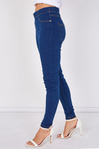 A person shown from the waist down wearing dark blue skinny jeans and white heeled sandals against a plain light gray studio background.