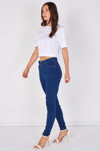 A woman modeling dark blue skinny jeans, paired with a white cropped t-shirt and white heeled sandals, standing against a plain white studio background.