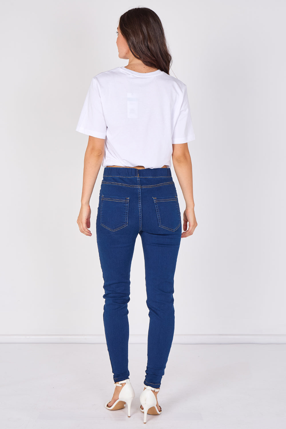 A woman viewed from behind wearing dark blue skinny jeans, a white t-shirt, and white high-heeled sandals against a plain white studio background.