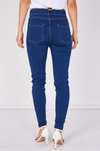 Rear view of a person wearing dark blue skinny jeans with white heeled sandals against a plain light gray studio background.