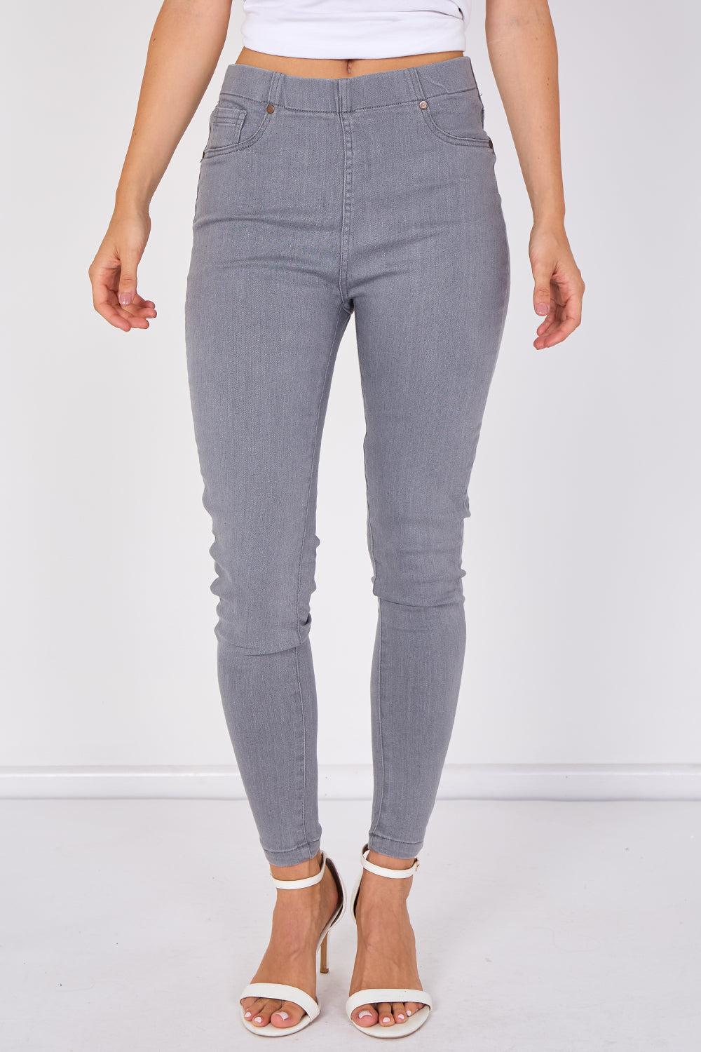 A model wearing high-waisted, light gray skinny jeans with a white crop top and white heeled sandals, shown from the waist down against a white studio background.