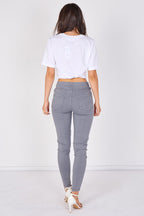 A woman viewed from behind wearing a white cropped T-shirt, gray skinny jeans, and white high-heeled sandals against a plain white studio background.