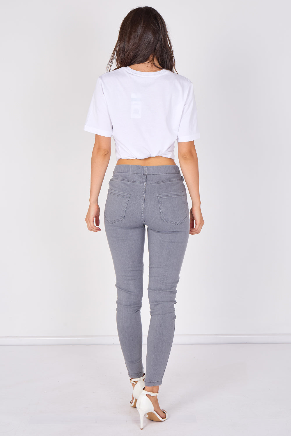A woman viewed from behind wearing a white cropped T-shirt, gray skinny jeans, and white high-heeled sandals against a plain white studio background.
