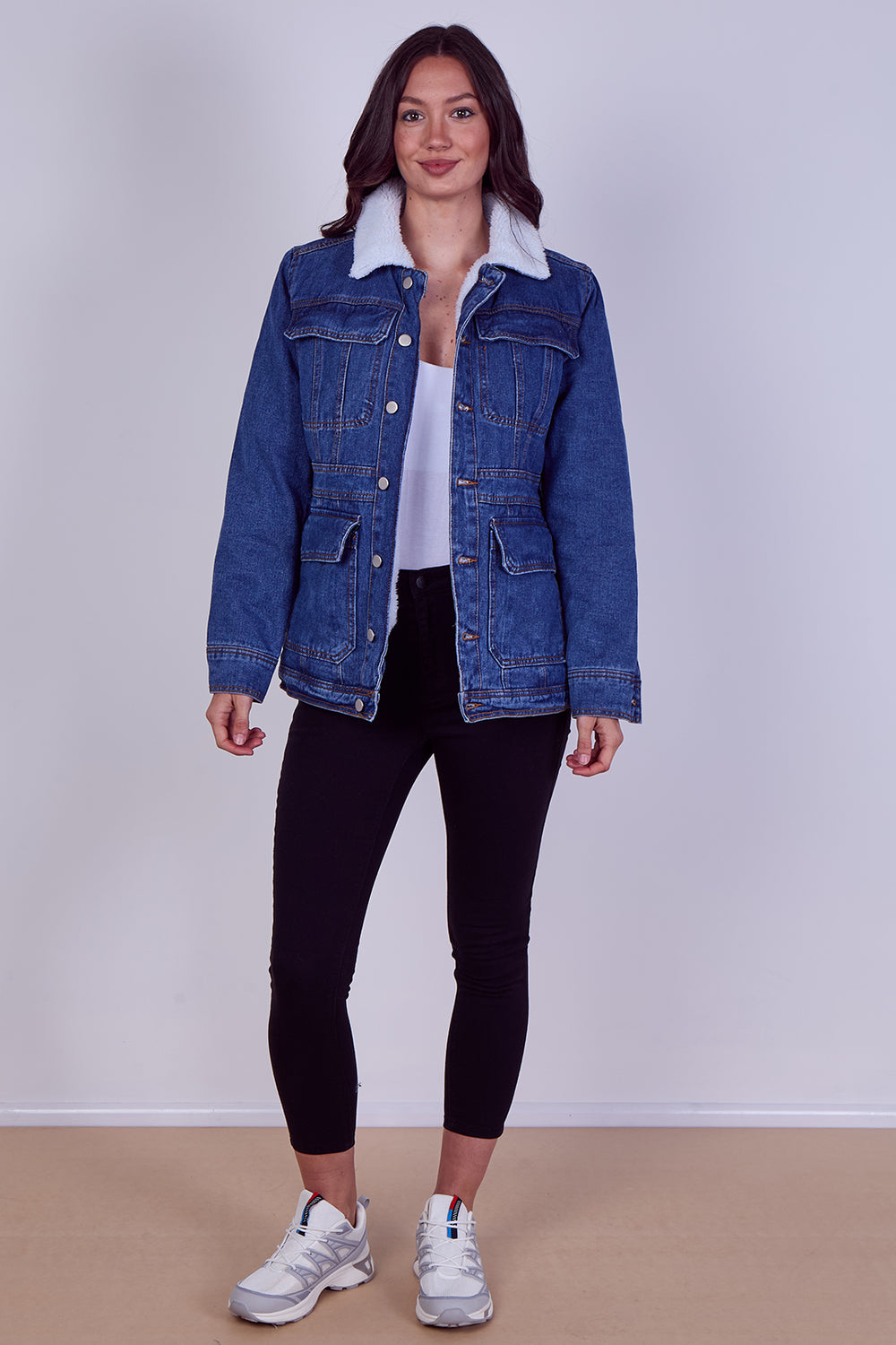 A woman modeling a blue denim jacket with a white sherpa collar, worn over a white top and black leggings, standing against a light gray studio wall.