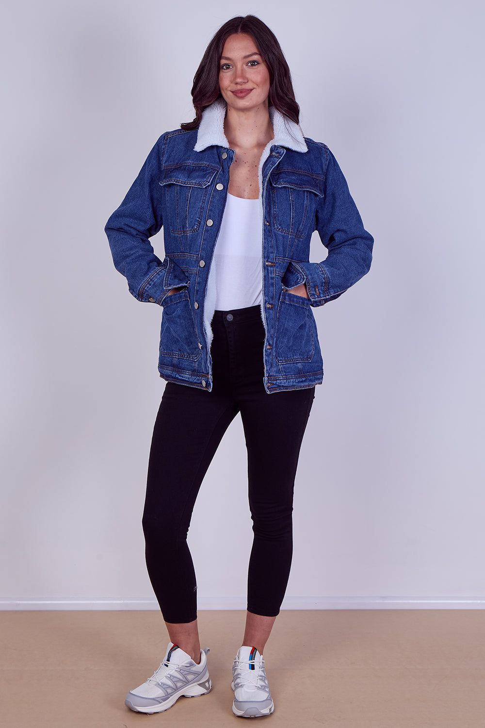 A woman modeling a blue denim jacket with a white sherpa collar, worn over a white tank top, black leggings, and white sneakers, standing against a light gray wall and beige floor.