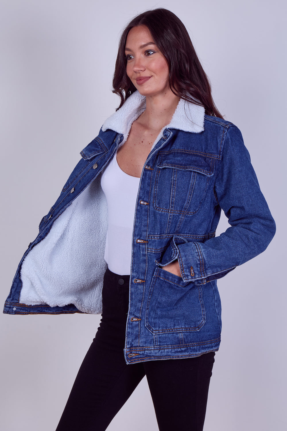 A woman modeling a blue denim jacket with a white sherpa lining, worn open over a white top and black pants, against a light gray studio background.