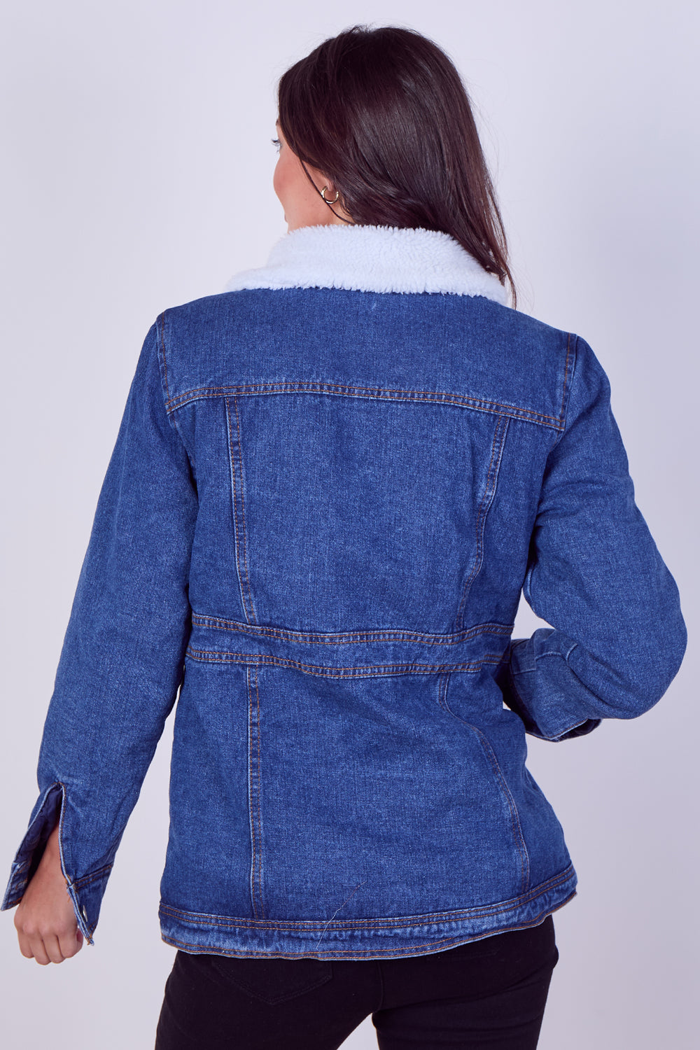 A woman shown from the back wearing a blue denim jacket with a white sherpa collar, paired with black pants, against a light gray studio background.