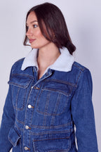 A woman wearing a blue denim jacket with a white sherpa fleece collar, shown from the chest up against a light gray studio background.