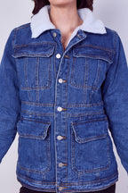 A blue denim jacket with a white sherpa fleece collar, featuring front button closure, chest flap pockets, and lower patch pockets, shown on a model against a light gray background.