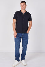 A man modeling a short-sleeve button-up shirt in dark navy blue with vertical textured stripes, paired with blue jeans and white sneakers, standing against a plain white studio background.