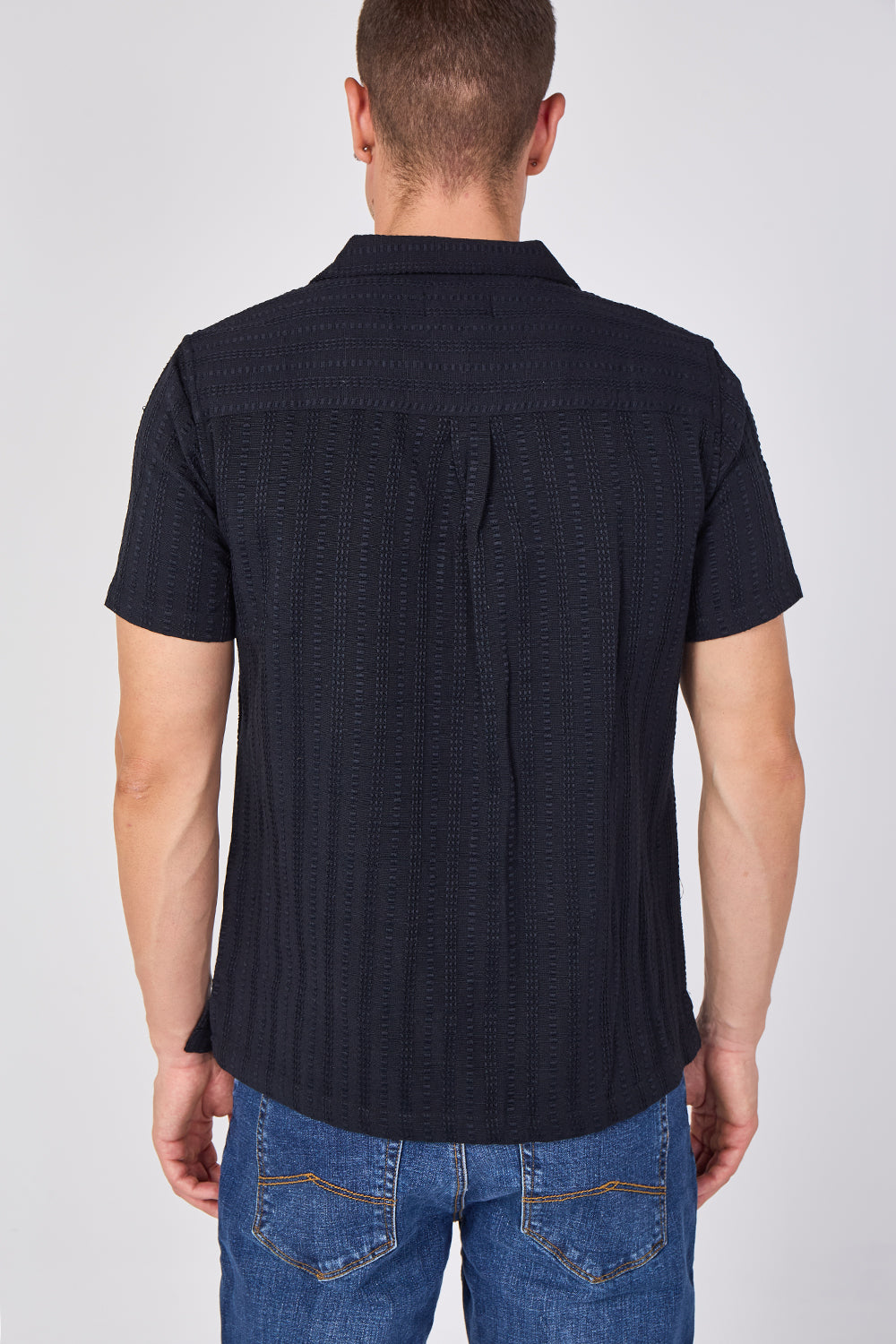 A man viewed from the back wearing a dark navy short-sleeve textured button-up shirt and blue jeans, set against a plain light gray studio background.