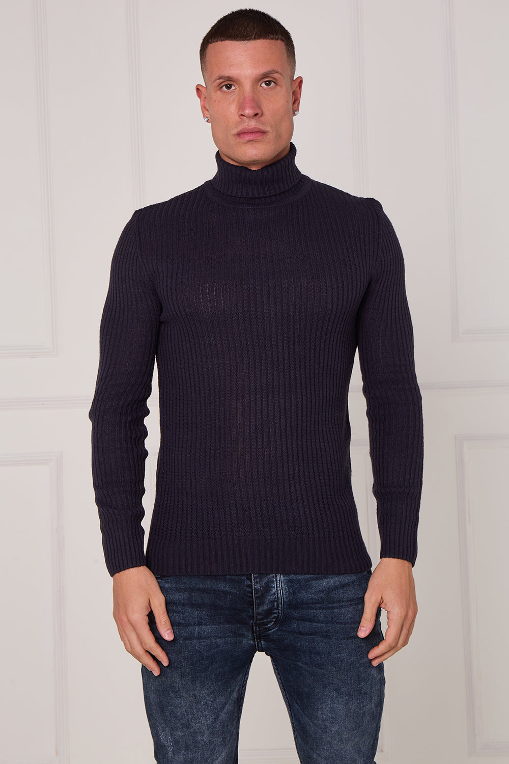 Man wearing a dark ribbed turtleneck sweater and jeans against a white wall. Main image