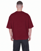 A man viewed from the back wearing an oversized short-sleeve crewneck t-shirt in burgundy and dark cargo pants, shot against a solid white background.