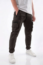 A person wearing dark olive cargo jogger pants with multiple zippered pockets, paired with a white t-shirt and white sneakers, shown from the waist down on a white studio background.