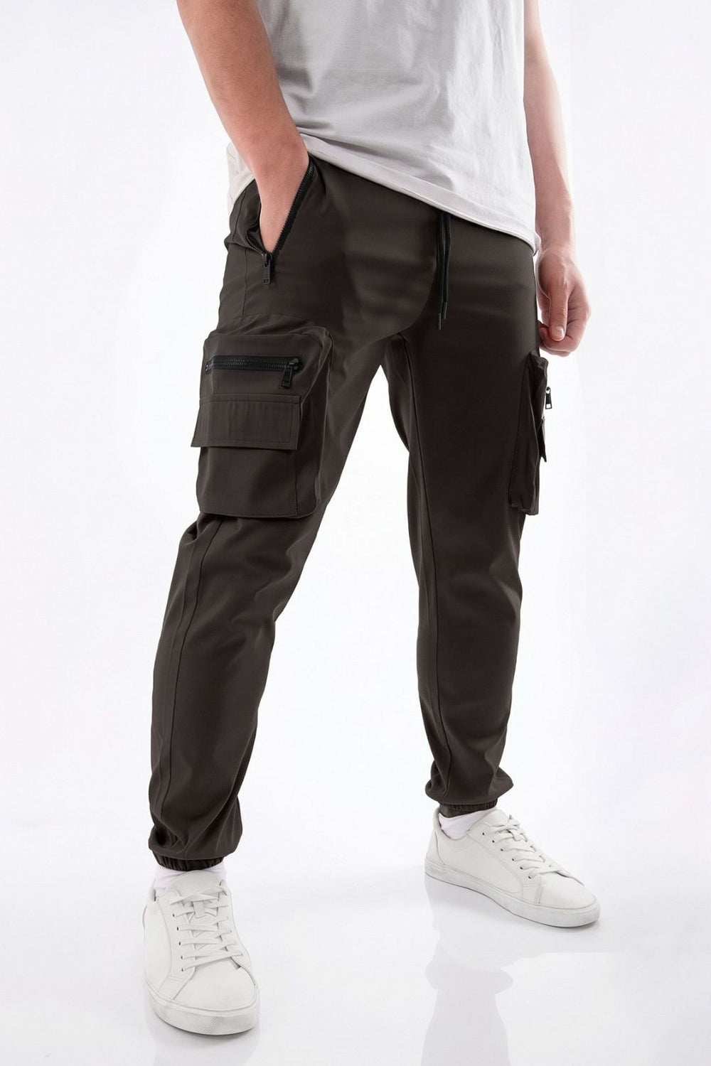 A person wearing dark olive cargo jogger pants with multiple zippered pockets, paired with a white t-shirt and white sneakers, shown from the waist down on a white studio background.
