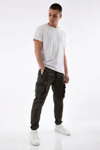 A man modeling dark olive green cargo pants, paired with a white t-shirt and white sneakers, standing against a seamless white studio background.