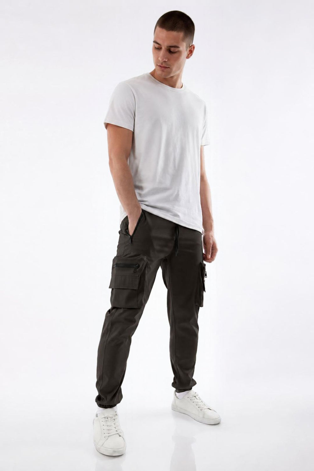 A man modeling dark olive green cargo pants, paired with a white t-shirt and white sneakers, standing against a seamless white studio background.